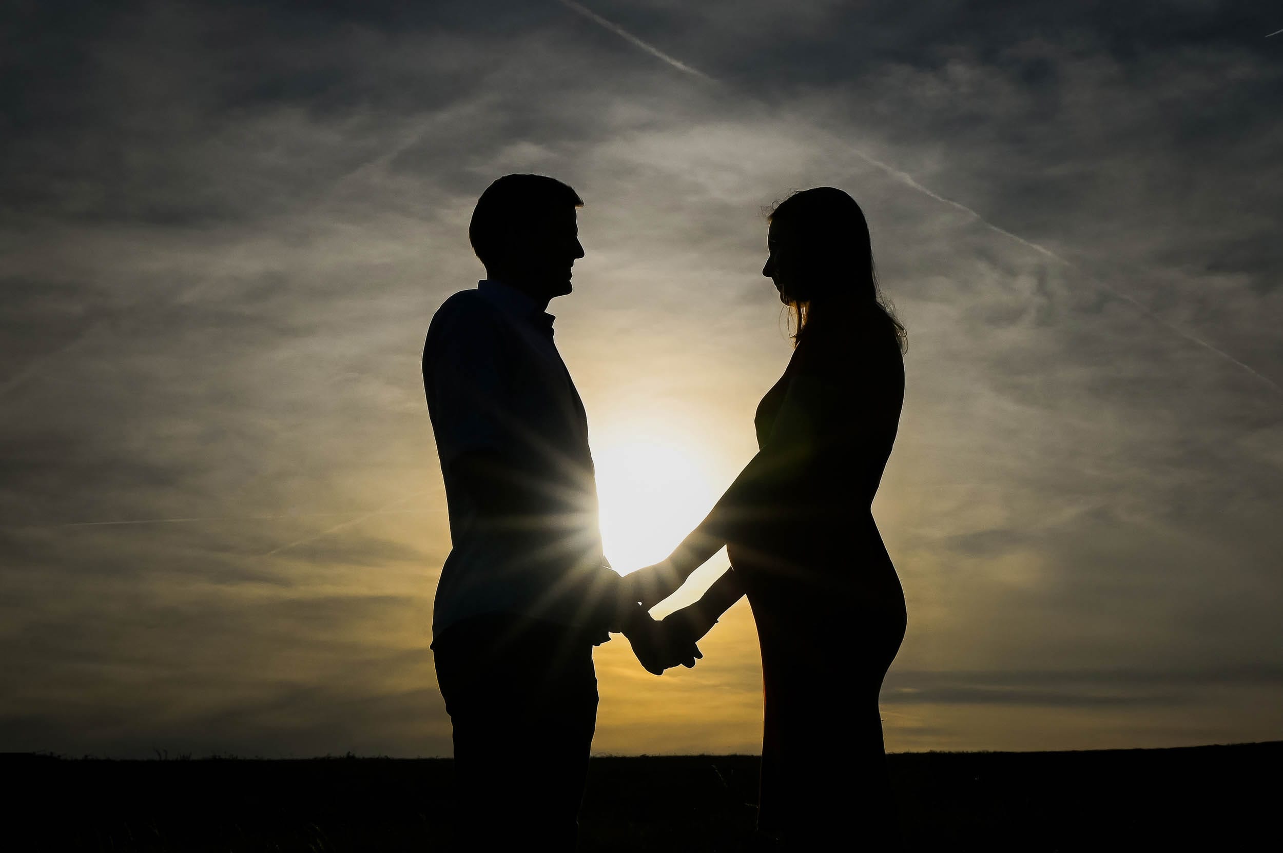 Silhoueta páru při těhotenském focení. Jemný moment očekávání zachycený při západu slunce.
